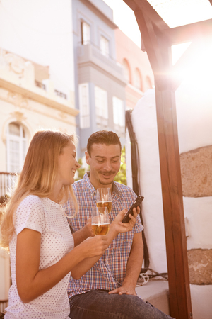 Relaxed smiling couple sitting on windowsill with beers and looking at a cellphone the blond girl is holdingの写真素材