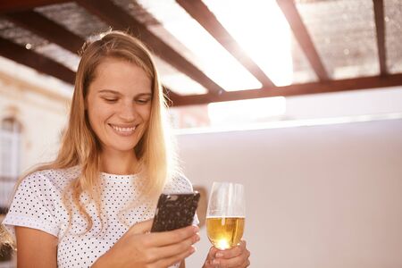 Beautiful blond with a toothy smile using her cellphone with her right hand while holding a beer in the other handの写真素材