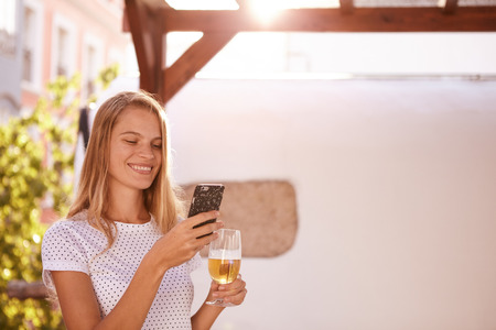 Pretty blond girl with a lovely smile looking at her cellphone while holding a beer in the other hand in overexposed sunlightの写真素材