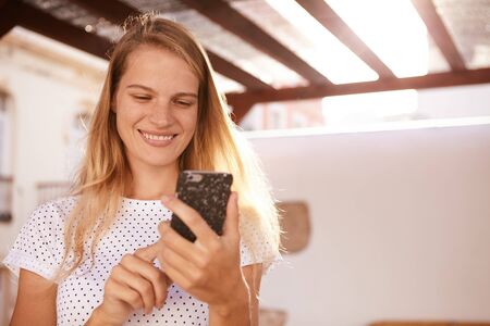 Happily smiling blond girl using her cellphone while looking at it and her long hair loose with the sun behind herの写真素材