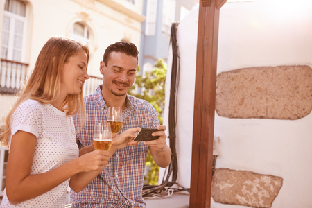 Happily smiling couple looking at cellphone that he is holding and she is pointing at while enjoying some beersの写真素材