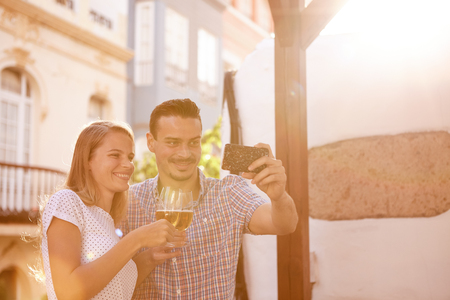 Happily smiling couple taking a selfie while raising their beer glasses and smiling happily in bright sunshineの写真素材