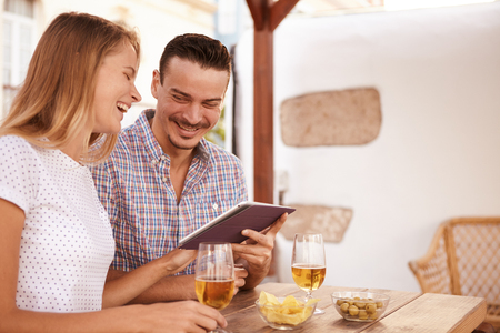 Goodlooking couple on a date laughing merrily while looking and pointing at a touchpad tablet with beers and snacks on the tableの写真素材