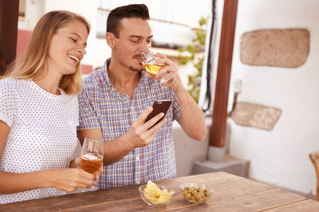 Young man sipping at his beer while showing his girlfriend something on his cellphone as she smiles a toothy smile with a beer in handの写真素材