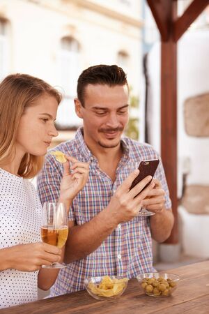 Young couple on a date looking at his cellphone while eating some snacks and drinking some beers in the bright afternoon sunlightの写真素材