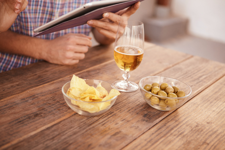 Glass bowls with crispy chips and green olives arranged with a beer glass on a rustic wooden table with relaxed hands holding a touchpadの写真素材