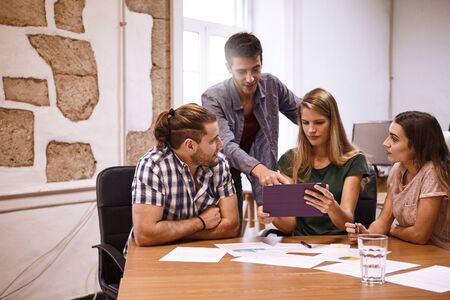 Team of four young professionals in an interesting and captivating strategic discussion in a room with a conference table strewn with notes and tablet pcの写真素材