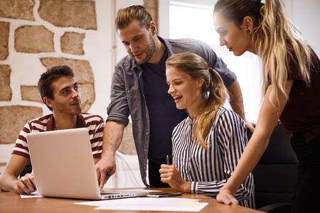Young business team sitting and standing around a laptop computer while one male is pointing at the keyboard of the laptopの写真素材