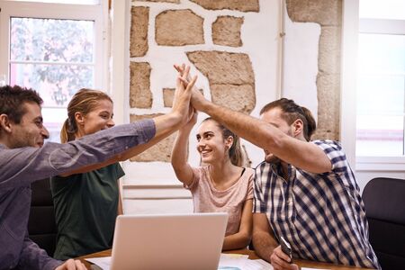 Team of four young business people gesturing high five to each other with big smiles to celebrate a breakthrough in their discussionsの写真素材