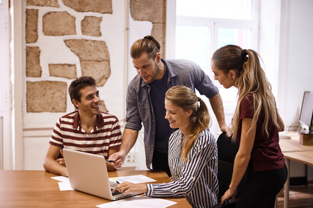 Young business team sitting and standing around a laptop computer while one male is pointing at the screen with his penの写真素材