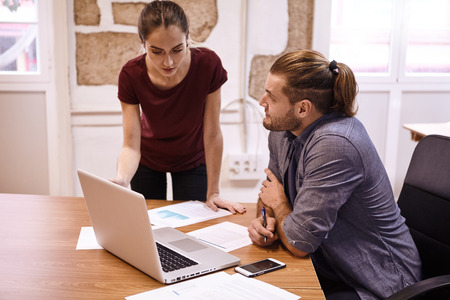 Young business woman instructing young man while standing next to him as she points at the laptop while he looks at herの写真素材