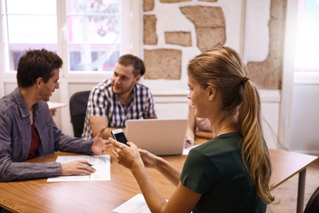 Young professional female sending message on the phone, with two young male professionals in the background in a meetingの写真素材