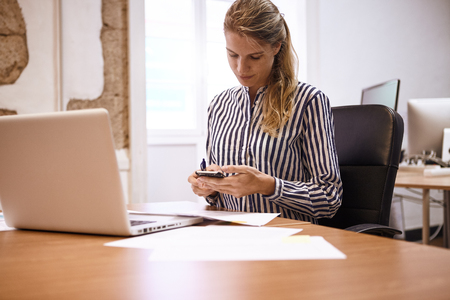 Pretty young business woman looking at her phone while sitting at her desk in the office with a laptop in front of herの写真素材