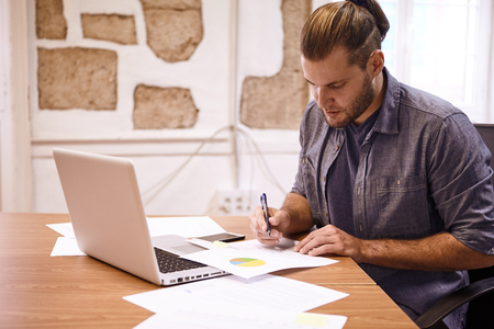 Businessman with tied back hair sitting at his desk with a laptop reading a chart in front of him with a pen in his handの写真素材