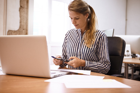 Young business woman making calculations on her phone while sitting at her desk with a laptop and various notes in front of herの写真素材