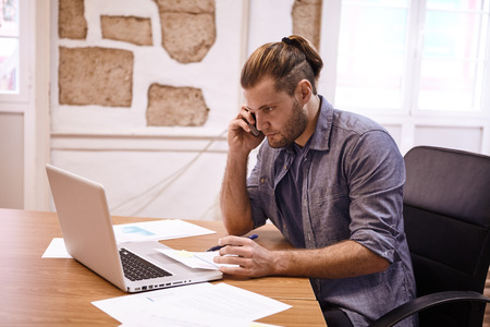 Young businessman in an office speaking on his phone while looking at his laptop with note paper in front of him and holding a penの写真素材