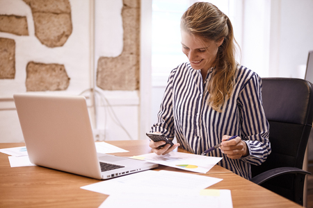 Young business woman smiling at her cellphone holding a pen in her left hand with a laptop in front of herの写真素材