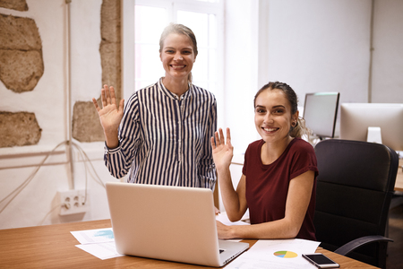 Two young business women smiling big toothy smiles and waving at the camera while sitting at a desk with a laptop and cellphoneの写真素材