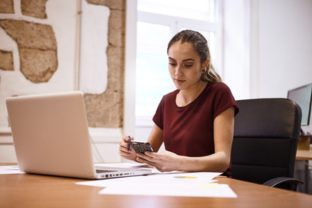 Intelligent young business woman at her desk with a laptop looking at her cellphone with a pen in the other hand looking thoughtfulの写真素材