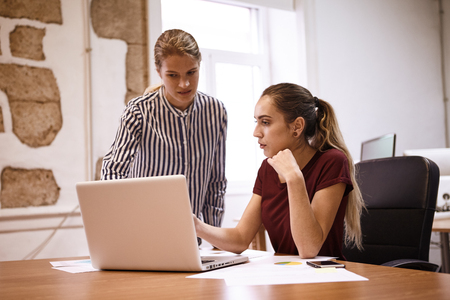 Two concerned business women looking and pointing worriedly at the laptop on the desk in front of them with a cell phoneの写真素材