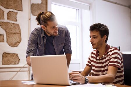 Young businessman being instructed by a male colleague while looking at his laptop on the desk in front of himの写真素材