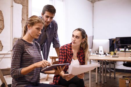 Team of three young business people looking slightly concerned while one lady is typing something on the tablet she is holdingの写真素材