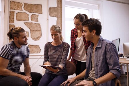 Young business team sitting and standing around in a semi circle all looking at the tablet in a lively discussionの写真素材