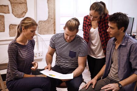 Young business team looking at pie charts while sitting and standing in a semi circle in a brightly...