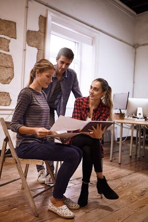 Two young ladies and a gentleman in a team building meeting in a brightly lit open plan office using notes and a tablet as toolsの写真素材
