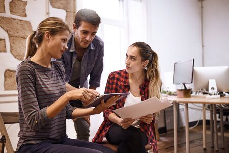 Concentrating team of three young business people pointing and looking at a tablet they are sharing in a very bright white officeの写真素材