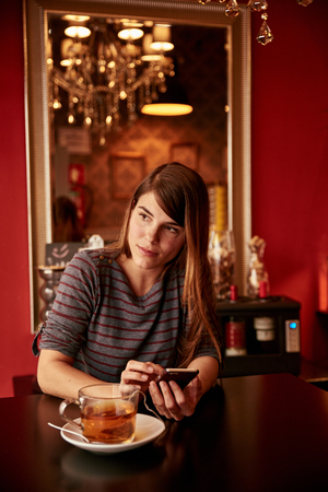 Pretty young lady texting while sitting at table wearing a gray and red striped top and her long hair loose, looking out the windowの写真素材
