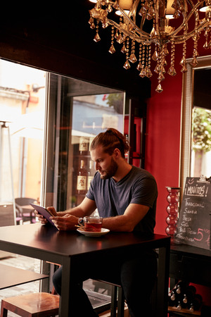 Handsome young lad sitting in a pub looking at his tablet with interest with a mirror and bar fridge behind himの写真素材