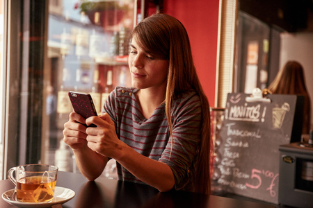 Cute young lady looking at her cell phone with interest and a secretive smile with the bar specials board behind herの写真素材