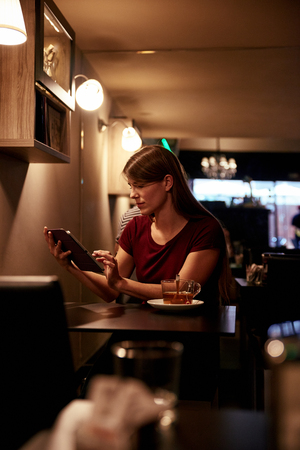 Beautiful young woman sitting at a table in a pub with a cup of tea using her tablet with her lovely long brown hair looseの写真素材