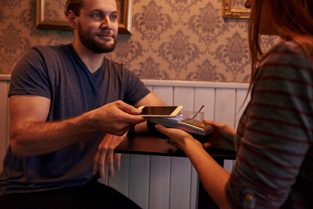Handsome man presenting his cell phone to lady with a paying machine in her hands while looking at her and smilingの写真素材