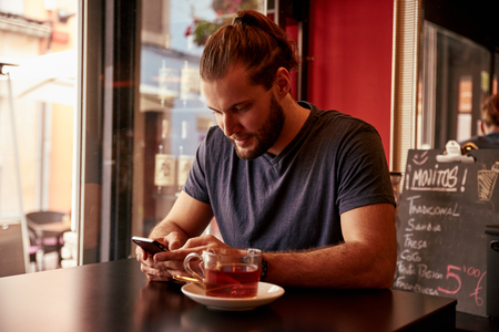 Smiling young man reading a cell phone message while sitting in a pub with a cup of tea on the table and a fridge behindの写真素材