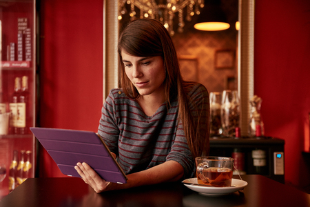 Beautiful young woman sitting in a bar looking at her tablet with a lovely smile in front of a gold framed mirrorの写真素材