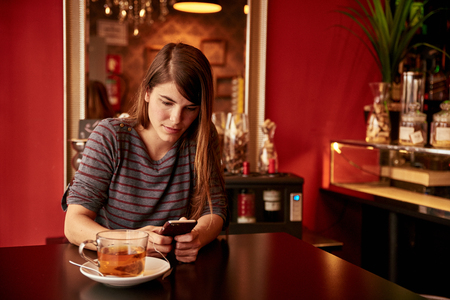 Pretty young lady looking very pensive while texting sitting at table wearing a gray and red striped top and her long hair looseの写真素材