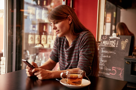 Pensive young lady looking at her cell phone while sitting in a bar with her long hair hanging loose and a cup of teaの写真素材