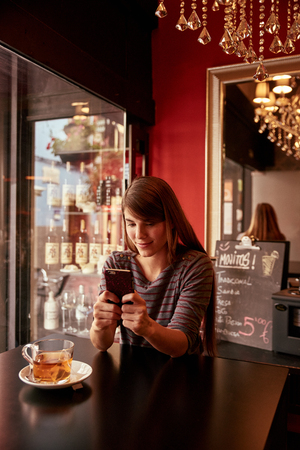 Pretty young lady sitting in a pub focused on her cell phone with a cup of tea on the table and a mirror behind herの写真素材