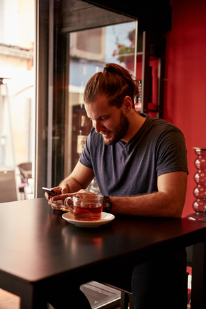 Young man with his long hair tied up sitting in a pub reading a message on his phone while wearing a blue t-shirtの写真素材