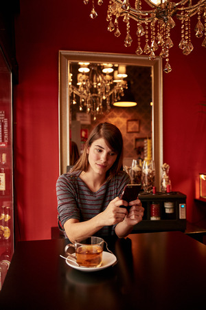 Cute young lady sitting at table sending a message on her cell phone wearing a gray and red striped top and her long hair looseの写真素材