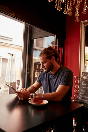 Interesting young man sitting in a pub looking at his tablet with his long hair tied up and a bar fridge behind himの写真素材