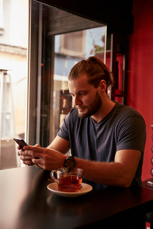 Young man with his hair tied in up texting on his phone while sitting in a pub with a cup of tea on the tableの写真素材