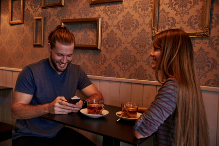 Dating young couple at a small table in a bar with big smiles while he is texting on his cell phoneの写真素材