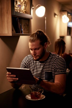 Goodlooking young man sitting at a table in a pub with a cup of tea using his touchpad while dressed in a striped t-shirtの写真素材