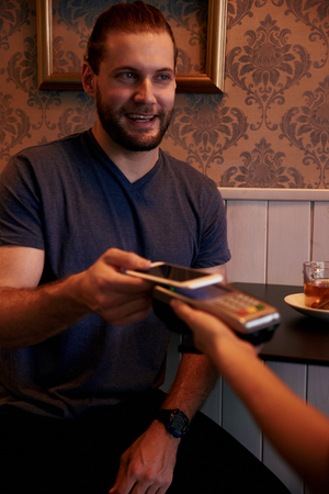 Handsome man offering his cell phone above the payment machine while looking at the person holding it with a toothy smileの写真素材