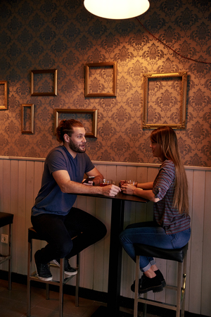 Happy young couple sitting on high bar stools at a small table in a pub while looking and talking with each otherの写真素材