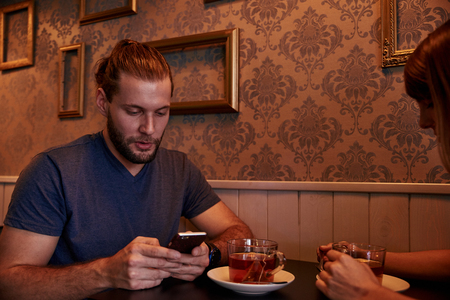 Young couple sitting together in a pub while he is texting on his cellphone she is looking at the teacup in her handsの写真素材