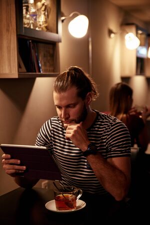 Goodlooking young man sitting at a table in a bar with a cup of tea using his touchpad while dressed in a striped t-shirtの写真素材
